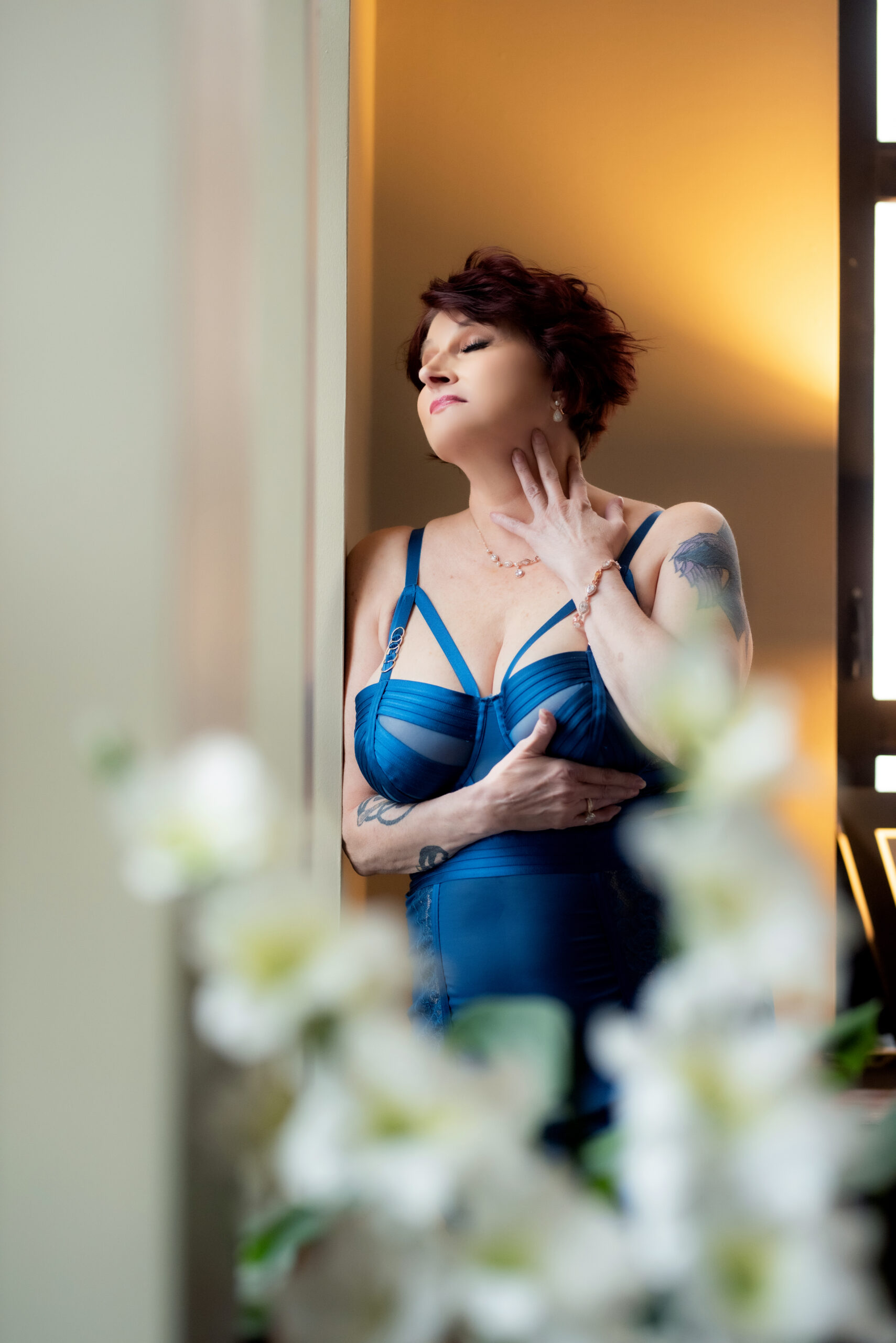 An older woman with short reddish-brown hair poses confidently in a blue lingerie set in the sun room hidden behind the beautiful monstera plant. The setting is the opulent Gatsby penthouse in Midtown, St. Louis, Missouri, owned by Josh Gould of Gould Holdings. The image was captured by Aloha Kelly of Love Exposed Boudoir.