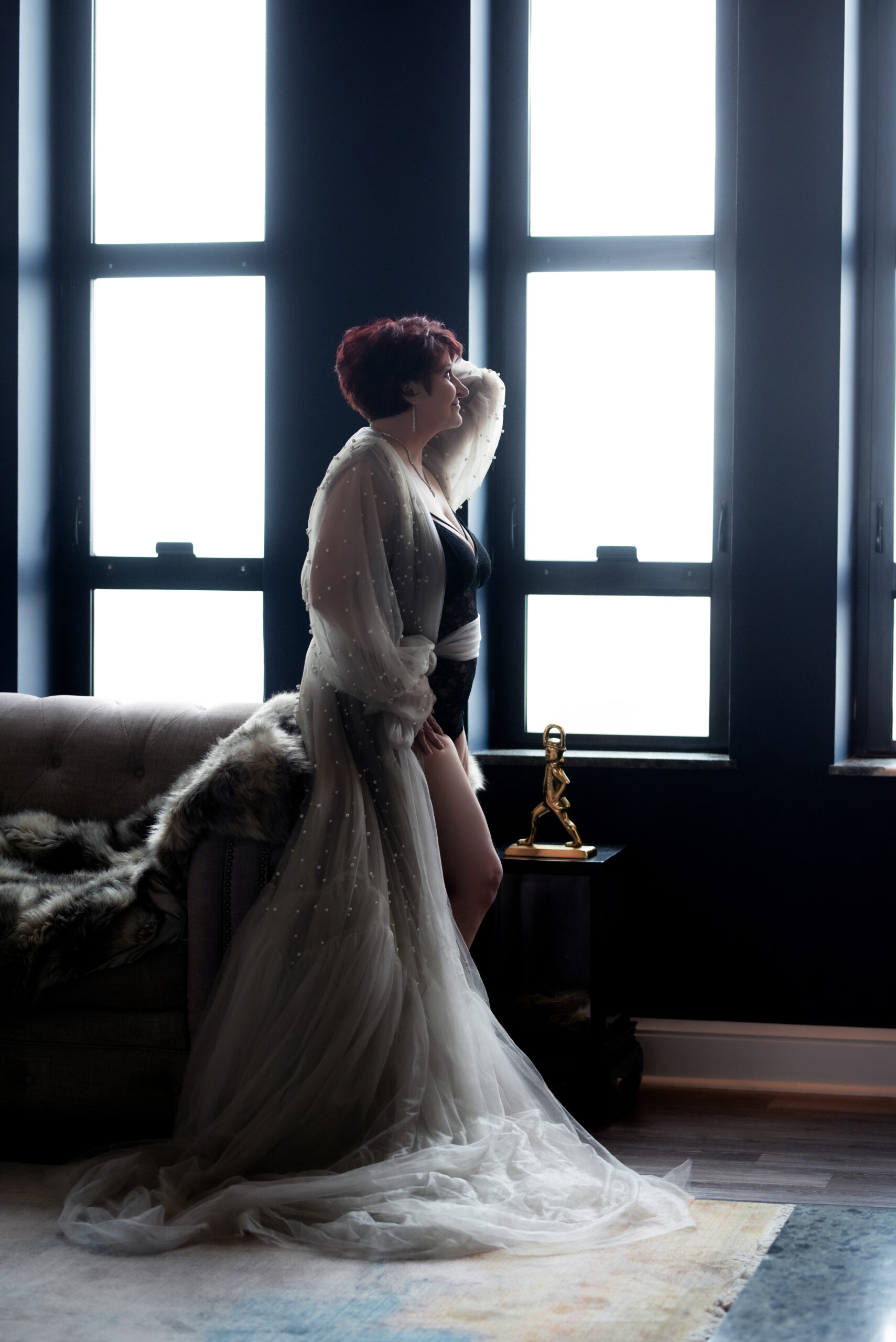 An older woman with short reddish-brown hair poses confidently in a gray pearl detail robe and a black lingerie set and high heels on the living room couch overlooking the view of downtown. The setting is the opulent Gatsby penthouse in Midtown, St. Louis, Missouri, owned by Josh Gould of Gould Holdings. The image was captured by Aloha Kelly of Love Exposed Boudoir.