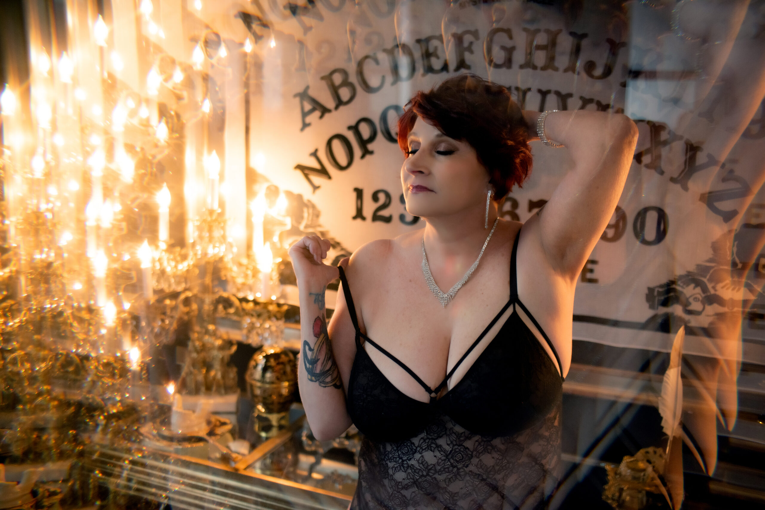 Lit by candle light, an older woman with short reddish-brown hair poses confidently in a black lingerie set in front of the iconic ouija board print in the gold and black master bedroom. The setting is the opulent Gatsby penthouse in Midtown, St. Louis, Missouri, owned by Josh Gould of Gould Holdings. The image was captured by Aloha Kelly of Love Exposed Boudoir.