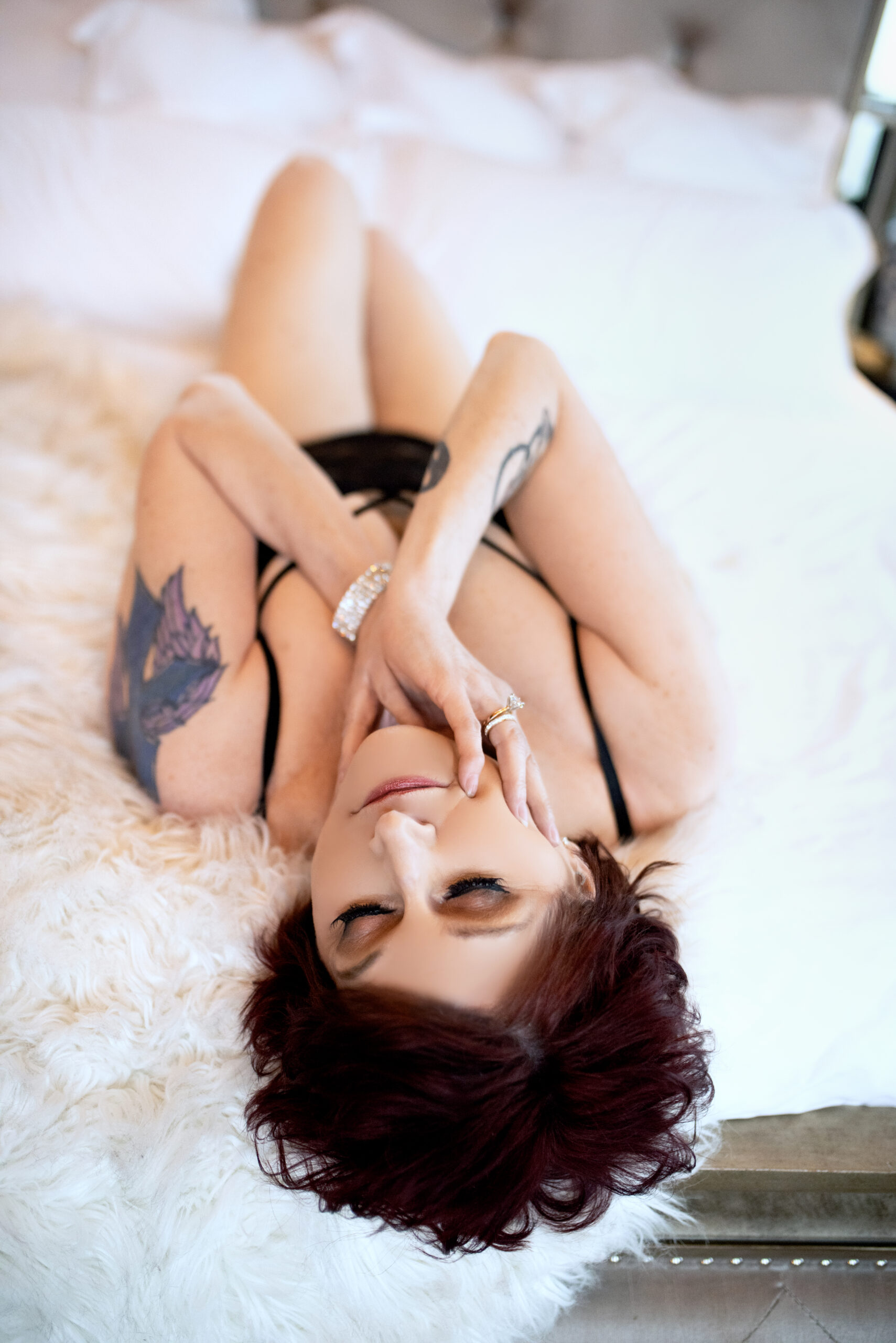 Lying on the gorgeous master bed adorned with a white fur blanket, an older woman with short reddish-brown hair poses confidently in a black lingerie set showing off her wedding ring. The setting is the opulent Gatsby penthouse in Midtown, St. Louis, Missouri, owned by Josh Gould of Gould Holdings. The image was captured by Aloha Kelly of Love Exposed Boudoir.